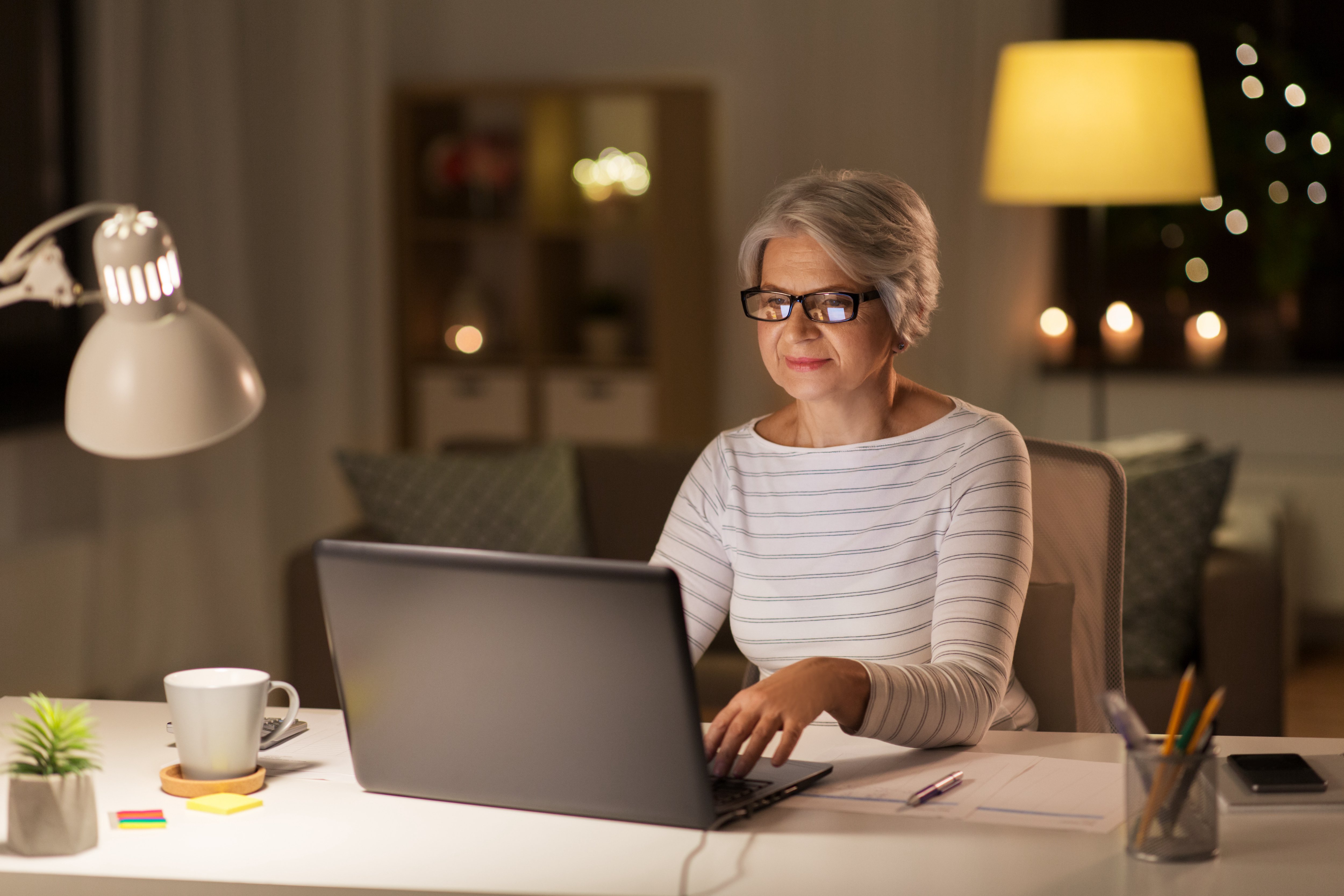 Older woman in glasses working on her laptop in a softly lit home office 